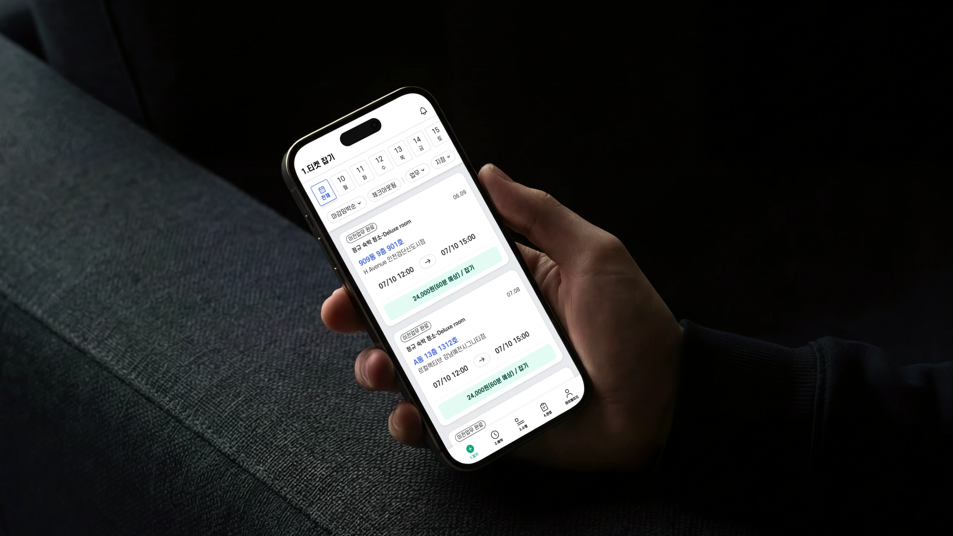 어두운 실내에서 손에 든 아이폰에 켜진 Keeper 앱 업무 리스트 — 날짜 스트립과 필터 칩 아래로 숙박 청소 업무 카드가 이어진다.